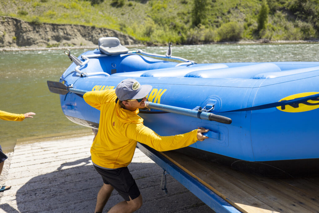A rafting guide in a yellow shirt takes a blue inflatable raft off a trailer.
